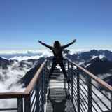 person standing on hand rails with arms wide open facing the mountains and clouds
